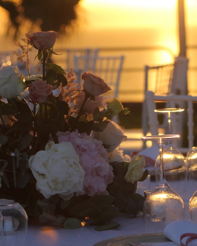 Tavola apparecchiata con centrotavola di rose e ortensie e calici, al tramonto.