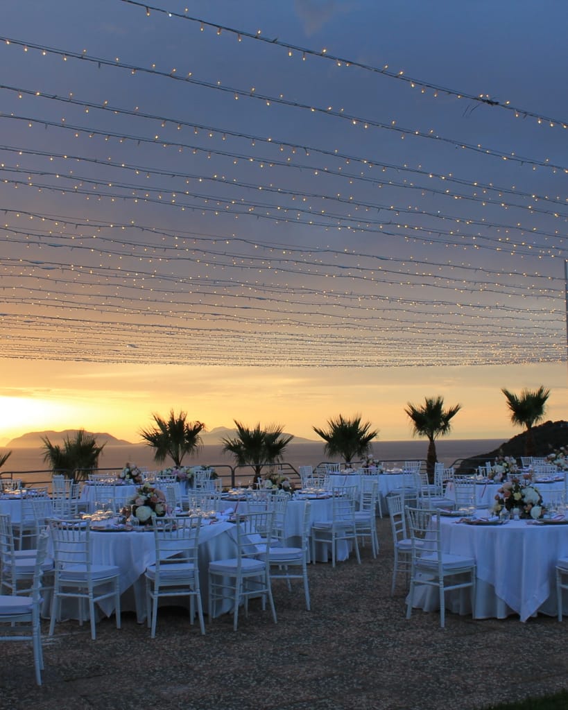 Tavoli apparecchiati sulla terrazza di Villa Hera sotto le luci a filo, al tramonto sul mare.