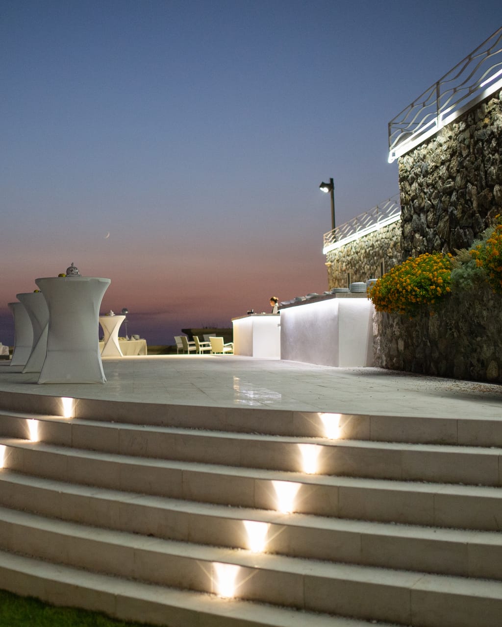 Scalinata curva illuminata e tavolini da aperitivo sulla terrazza di Villa Hera, all’ora blu.