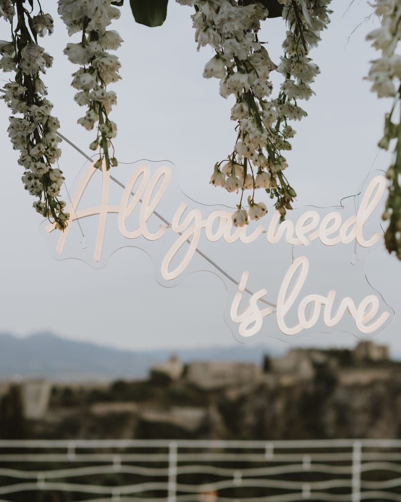 Scritta al neon "All you need is love" tra i grappoli di glicine; sullo sfondo, sfocato, il Castello di Milazzo.