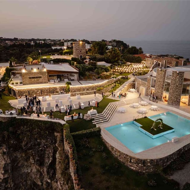 Veduta aerea di Villa Hera all’imbrunire, durante una festa: ospiti tra il prato e la piscina, con la Torre Saracena illuminata sullo sfondo.