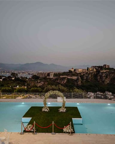 Piscina di Villa Hera con la piattaforma a prato e l’arco floreale, al crepuscolo, con il Castello di Milazzo sullo sfondo.